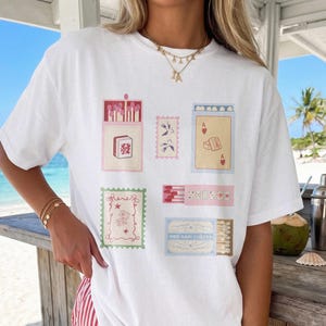 May include: A white short-sleeve t-shirt featuring a graphic print of various matchbox-style illustrations, including a mahjong tile, playing cards, swallows, and decorative stamps. The design incorporates soft pastel colors and small text details.