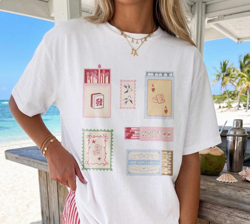 May include: A white short-sleeve t-shirt featuring a graphic print of various matchbox-style illustrations, including a mahjong tile, playing cards, swallows, and decorative stamps. The design incorporates soft pastel colors and small text details.