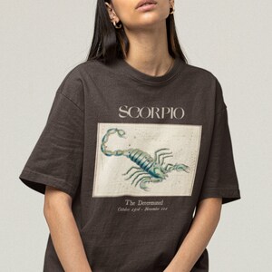 Op de afbeelding: Een bruin t-shirt met een grafische print van een schorpioen en de tekst "SCORPIO" en "The Determined, October 23rd - November 21st".