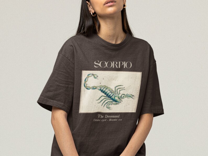 Op de afbeelding: Een bruin t-shirt met een grafische print van een schorpioen en de tekst "SCORPIO" en "The Determined, October 23rd - November 21st".