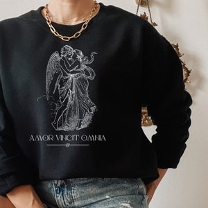 May include: Black sweatshirt with a white graphic of two figures embracing. The graphic is a stylized illustration of a winged figure with a halo, embracing another figure. The text "AMOR VINCIT OMNIA" is printed below the graphic.