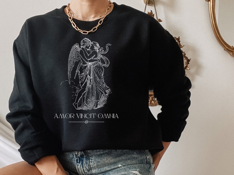 May include: Black sweatshirt with a white graphic of two figures embracing. The graphic is a stylized illustration of a winged figure with a halo, embracing another figure. The text "AMOR VINCIT OMNIA" is printed below the graphic.
