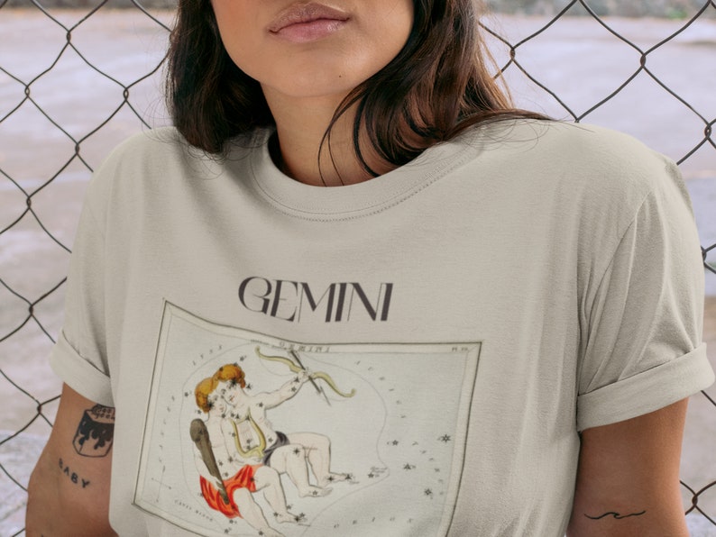 Pode incluir: Uma camiseta bege claro com uma ilustra&ccedil;&atilde;o vintage da constela&ccedil;&atilde;o de G&ecirc;meos e a palavra "GEMINI" impressa em preto.