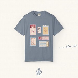 May include: A blue jean colored short-sleeve t-shirt featuring a graphic print of various rectangular matchbox designs in shades of red, pink, and cream. The shirt has a crew neckline and a centered arrangement of illustrated matchbox motifs.