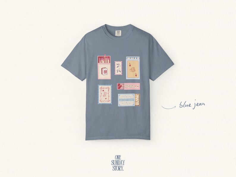 May include: A blue jean colored short-sleeve t-shirt featuring a graphic print of various rectangular matchbox designs in shades of red, pink, and cream. The shirt has a crew neckline and a centered arrangement of illustrated matchbox motifs.