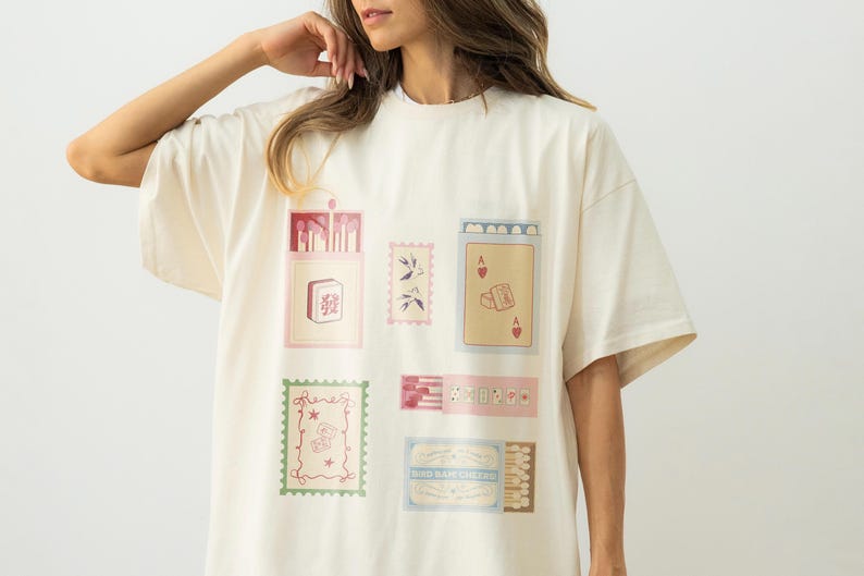 May include: An off-white short-sleeve t-shirt featuring a graphic print of various rectangular illustrations, including a mahjong tile, a playing card with an ace of hearts, postage stamps, and matchboxes with detailed line art and soft colors.
