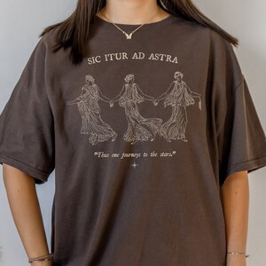 May include: A brown t-shirt with a white graphic print of three women holding hands. The text "Sic Itur Ad Astra" is printed above the graphic and the text "Thus one journeys to the stars." is printed below the graphic.