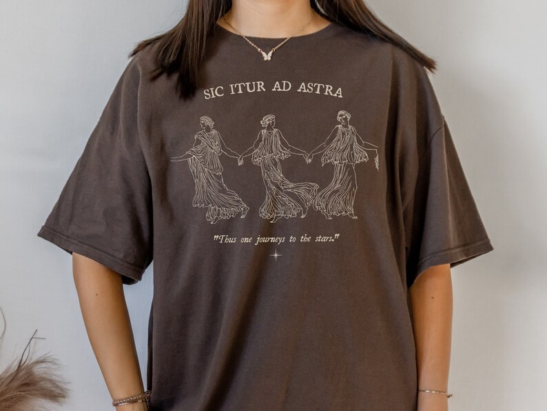 May include: A brown t-shirt with a white graphic print of three women holding hands. The text "Sic Itur Ad Astra" is printed above the graphic and the text "Thus one journeys to the stars." is printed below the graphic.