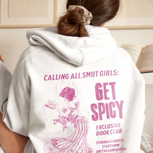 May include: A white hoodie with a pink graphic print that says "Calling all smut girls: Get Spicy" and "Exclusive Book Club" with a pink illustration of a woman reading a book. The text also says "111 Morally Greyst. Starts 8PM Only Villain Lovers!"