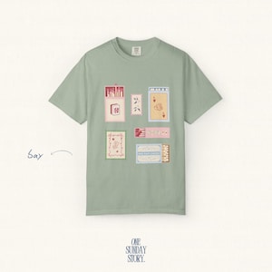 May include: A sage green short-sleeve t-shirt featuring a graphic print of various rectangular matchbox-style illustrations in shades of pink, red, and blue. The design includes small floral motifs and playing card imagery. The text "ONE SUNDAY STORY." is printed at the bottom.