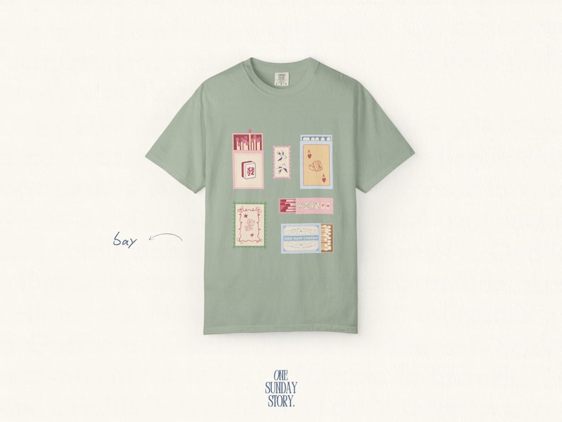 May include: A sage green short-sleeve t-shirt featuring a graphic print of various rectangular matchbox-style illustrations in shades of pink, red, and blue. The design includes small floral motifs and playing card imagery. The text "ONE SUNDAY STORY." is printed at the bottom.