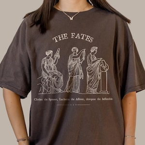 May include: A dark brown t-shirt with a white graphic print of three figures representing the Fates. The text "THE FATES" is printed above the figures. Below the figures, the text reads "Clotho the Spinner, Lachesis the Allotter, Atropos the Inflexible."