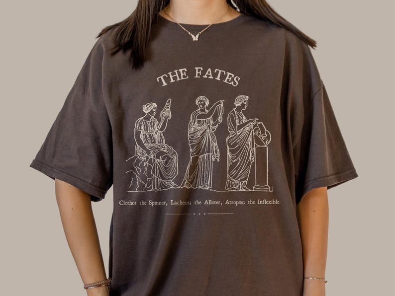 May include: A dark brown t-shirt with a white graphic print of three figures representing the Fates. The text "THE FATES" is printed above the figures. Below the figures, the text reads "Clotho the Spinner, Lachesis the Allotter, Atropos the Inflexible."