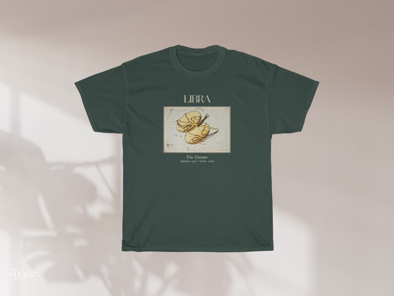 May include: Dark green t-shirt featuring the word "LIBRA" above a vintage-style illustration of the Libra zodiac symbol. The illustration is in shades of yellow and brown. Below the illustration is the text "The Charmer September 23rd - October 22nd."