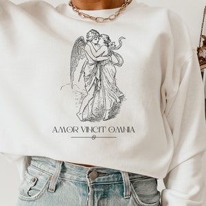 May include: White sweatshirt with a black and white illustration of a winged cupid embracing a woman. The text "AMOR VINCIT OMNIA" is printed below the illustration.