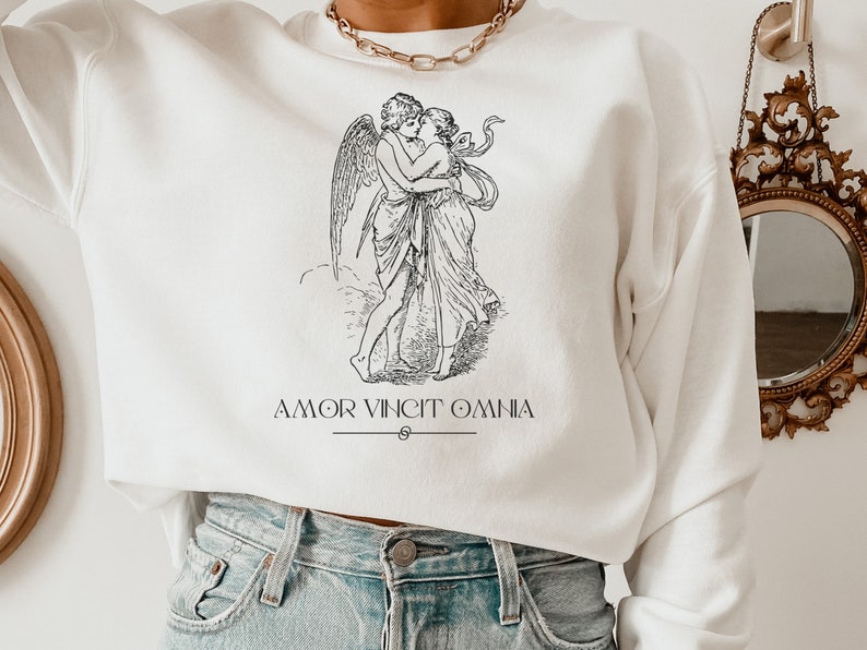 May include: White sweatshirt with a black and white illustration of a winged cupid embracing a woman. The text "AMOR VINCIT OMNIA" is printed below the illustration.