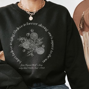 May include: Black sweatshirt with a white graphic print of a floral bouquet and a quote from Virginia Woolf's diary: "I don't believe in aging. I believe in forever altering one's aspect to the sun." The quote is from an entry dated October 2nd, 1932.