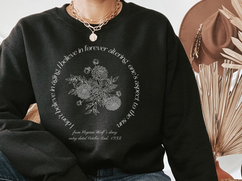 May include: Black sweatshirt with a white graphic print of a floral bouquet and a quote from Virginia Woolf's diary: "I don't believe in aging. I believe in forever altering one's aspect to the sun." The quote is from an entry dated October 2nd, 1932.