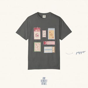 May include: A charcoal gray short-sleeve t-shirt featuring a graphic print of various matchboxes and postage stamps in shades of pink, blue, and cream. The word pepper is written in cursive to the right of the shirt with an arrow pointing to it.