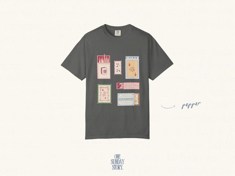 May include: A charcoal gray short-sleeve t-shirt featuring a graphic print of various matchboxes and postage stamps in shades of pink, blue, and cream. The word pepper is written in cursive to the right of the shirt with an arrow pointing to it.