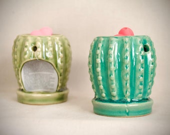 May include: Two ceramic cactus-shaped oil warmers. One is light green, the other is teal. Both have a pink top and a hole for the oil. The warmers have a textured surface and are on a small base.