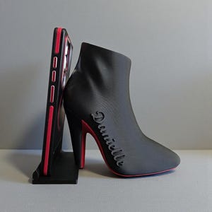 Op de afbeelding: Een zwarte telefoonstandaard in de vorm van een high-heeled laars met een rode zool en hak. Het woord "Danielle" staat op de zijkant van de laars. Een zwarte telefoonhoes met rode accenten staat in de standaard.