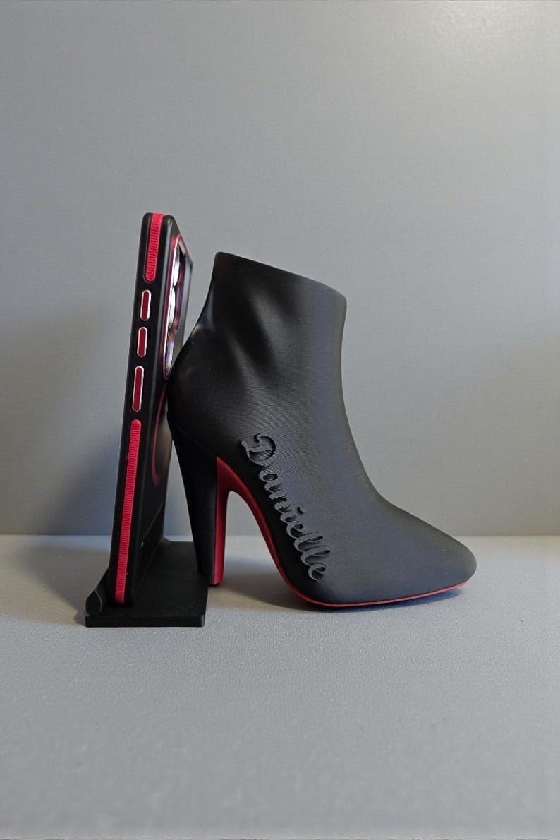 Op de afbeelding: Een zwarte telefoonstandaard in de vorm van een high-heeled laars met een rode zool en hak. Het woord "Danielle" staat op de zijkant van de laars. Een zwarte telefoonhoes met rode accenten staat in de standaard.