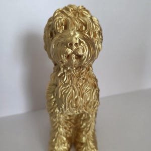 May include: A metallic gold-colored figurine of a curly-haired dog, likely a doodle breed, standing in a forward-facing position. The sculpture features detailed, textured fur patterns across its entire body, including the head, ears, and legs.