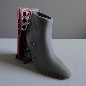 Op de afbeelding: Een zwart laarzenvormig object met de naam "Danielle" in cursief schrift. De laars heeft een rode zool en staat naast een zwart-rode telefoonhoes. De telefoonhoes heeft goudkleurige cameralenzen.