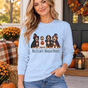 May include: A light blue long-sleeve shirt featuring a graphic of four dogs in Halloween costumes, including a witch, a ghost, a bat, and a wizard. The text below the dogs reads "BIG FLUFF, BIGGER BOOS" in a black, sans-serif font.