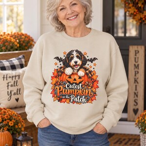 May include: A beige crewneck sweatshirt featuring a graphic print of a Bernedoodle puppy sitting inside a carved jack-o'-lantern pumpkin. The design includes autumn leaves, small bats, and the text "Cutest Pumpkin in the Patch" in orange.