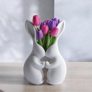 May include: A white ceramic vase shaped like two embracing rabbits, holding hands. The vase is filled with vibrant pink and purple tulips with green stems and leaves. The bunny ears form the vase's opening.