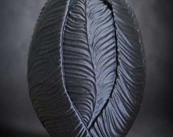 May include: A dark gray, textured, oval-shaped decorative object. The object has a petal-like design with vertical, wavy lines. The overall shape resembles a closed flower bud. The background is a dark gray.