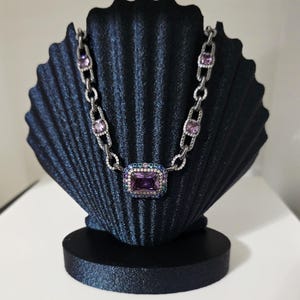 Puede incluir: Un collar plateado con un gran colgante rectangular de piedra preciosa púrpura y piedras preciosas rosas más pequeñas. El collar se exhibe en un expositor de joyas negro en forma de concha.