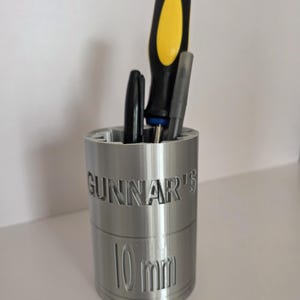 May include: A silver cylindrical desk organizer shaped like a large socket tool, featuring the embossed text "GUNNAR'S" and "10 mm" on the side. The container holds a black and yellow screwdriver, a black pen, and a grey pen in its open top.