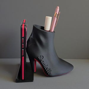 Op de afbeelding: Een zwarte bureau-organizer in de vorm van een hoge haklaars met een rode zool en hak. De laars bevat een roze pen en een witte tube. Naast de laars staat een zwarte telefoonhoes met rode accenten.