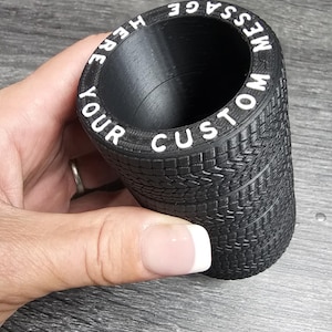 Pode incluir: Recipiente cilíndrico preto com um padrão de banda de rodagem de pneu. O recipiente tem letras brancas ao redor da parte superior que dizem "WRITE YOUR CUSTOM MESSAGE HERE".