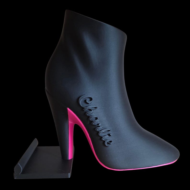 Op de afbeelding: Zwarte high-heeled laars met roze zool en hak, met de naam "Charlie" in cursief schrift. De laars staat op een zwarte standaard tegen een zwarte achtergrond. Het ontwerp is strak en modern.