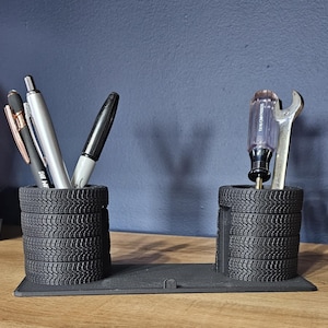 May include: Black desk organizer with two tire-shaped pen holders. One holds pens, the other a screwdriver and wrench. The organizer is on a black base, perfect for a mechanic's desk or automotive enthusiast.