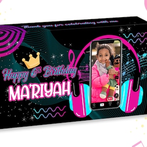 May include: A black and pink Skittles candy box with a personalized birthday message for Mariyah. The box features a design with a phone screen, headphones, musical notes, and a crown. The phone screen shows a photo of a girl with a pink shirt and a stuffed animal.