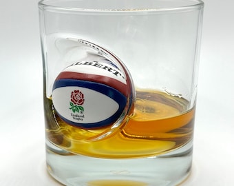 May include: A clear glass tumbler with a brown liquid and a white, red, and blue rugby ball inside. The ball has the words "Gilbert" and "England Rugby" printed on it.