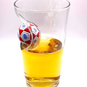 May include: A clear glass pint glass featuring a molded indentation on the side that holds a small, red and white soccer ball with the blue England national team crest printed on the panels. The glass is partially filled with a golden liquid.