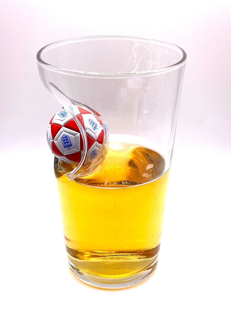 May include: A clear glass pint glass featuring a molded indentation on the side that holds a small, red and white soccer ball with the blue England national team crest printed on the panels. The glass is partially filled with a golden liquid.