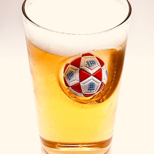 May include: A clear glass filled with golden beer and a thick white foam head. Embedded into the side of the glass is a three-dimensional soccer ball featuring red and white panels with blue crests printed on the white sections of the ball.