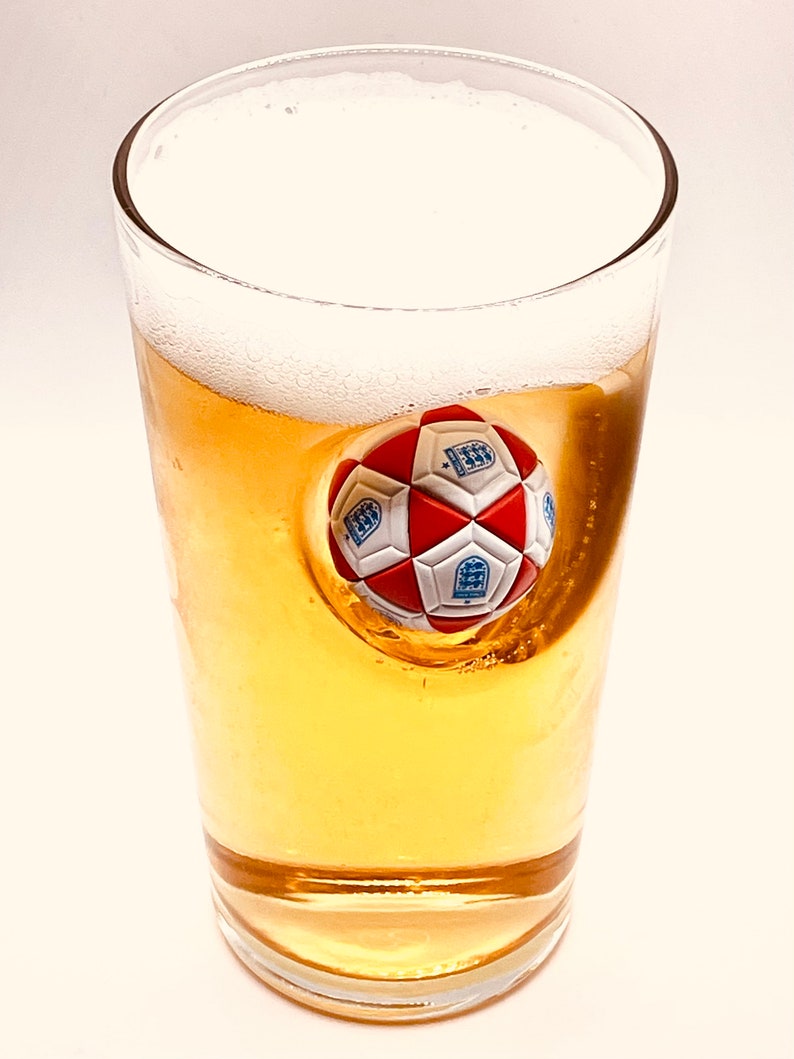 May include: A clear glass filled with golden beer and a thick white foam head. Embedded into the side of the glass is a three-dimensional soccer ball featuring red and white panels with blue crests printed on the white sections of the ball.