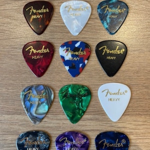 May include: Twelve guitar picks with the Fender logo and the word "Heavy" printed on them. The picks are in various colors and patterns, including black, white, red, blue, green, and purple.
