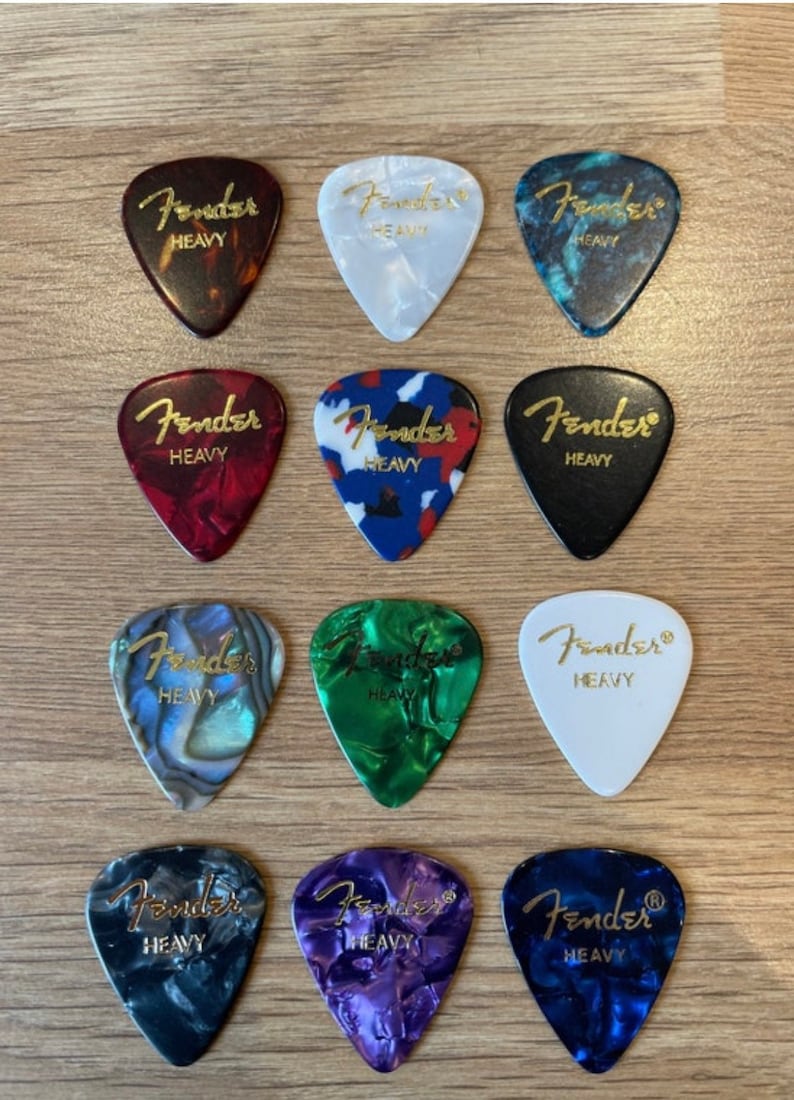May include: Twelve guitar picks with the Fender logo and the word "Heavy" printed on them. The picks are in various colors and patterns, including black, white, red, blue, green, and purple.