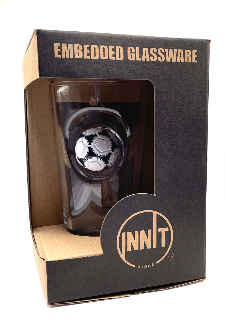May include: A clear glass tumbler featuring an embedded black and white soccer ball design, housed in a black cardboard box labeled EMBEDDED GLASSWARE and INNIT STORE. The glass is visible through a rectangular cutout in the front of the packaging.