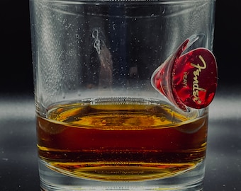 Puede incluir: Un vaso de vidrio transparente con un líquido marrón en su interior. Una púa de guitarra Fender roja está unida al lado del vaso.