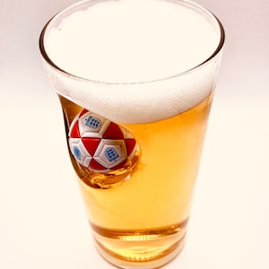 May include: A clear glass pint beer glass filled with golden liquid and a thick white foam head. A three-dimensional red and white soccer ball with blue England national team crests is embedded into the side of the glass, protruding inward.